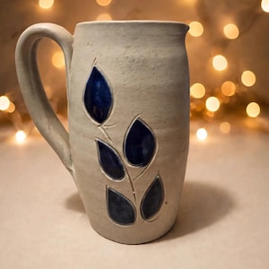 May include: A cylindrical ceramic pitcher with a matte off-white finish and a single handle. The exterior features a carved stem design with five glossy, deep blue glazed leaves arranged in a vertical pattern on the front surface.