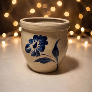 May include: A beige stoneware crock featuring a hand-painted dark blue floral design on the front. The vessel has a wide, open top with a thick, rounded rim and a tapered body that narrows toward the base, showcasing a smooth, glazed finish.