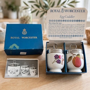 Puede incluir: Un juego de cocedores de huevos Royal Worcester en una caja azul con tapa que incluye instrucciones. Se incluyen dos cocedores de porcelana blanca con tapas metálicas y anillas, uno con diseño de moras y otro con un melocotón.