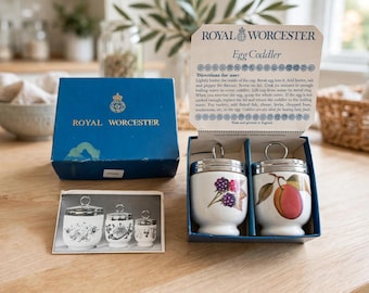 Puede incluir: Un juego de cocedores de huevos Royal Worcester en una caja azul con tapa que incluye instrucciones. Se incluyen dos cocedores de porcelana blanca con tapas metálicas y anillas, uno con diseño de moras y otro con un melocotón.