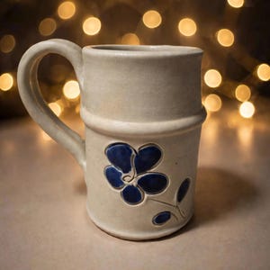 May include: A cream-colored ceramic mug with a cylindrical body and a prominent handle. The mug features a raised, hand-painted dark blue flower design with five petals and a small leaf on the lower section of the exterior surface.