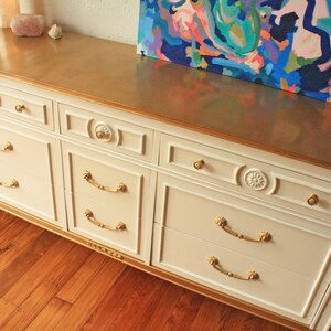 May include: A white dresser with gold trim and six drawers. The dresser has gold handles and decorative details on the drawers. The dresser is sitting on a wooden floor.