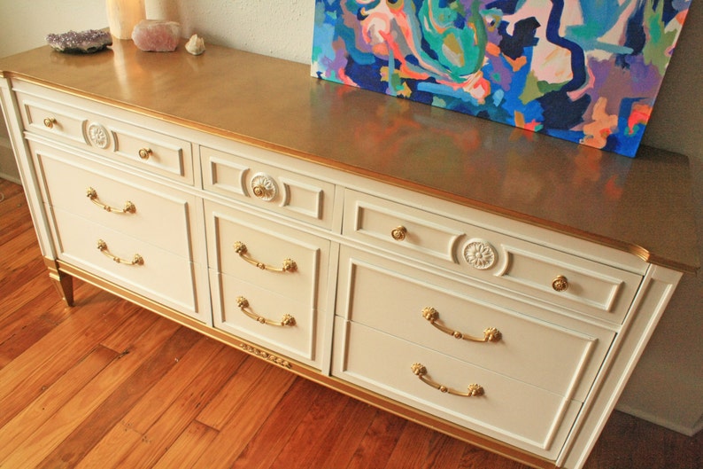 May include: A white dresser with gold trim and six drawers. The dresser has gold handles and decorative details on the drawers. The dresser is sitting on a wooden floor.