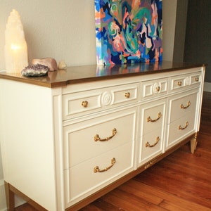 May include: A white dresser with gold accents and six drawers. The dresser has gold handles and decorative details. The dresser is sitting on a wooden floor. An abstract painting hangs on the wall behind the dresser.