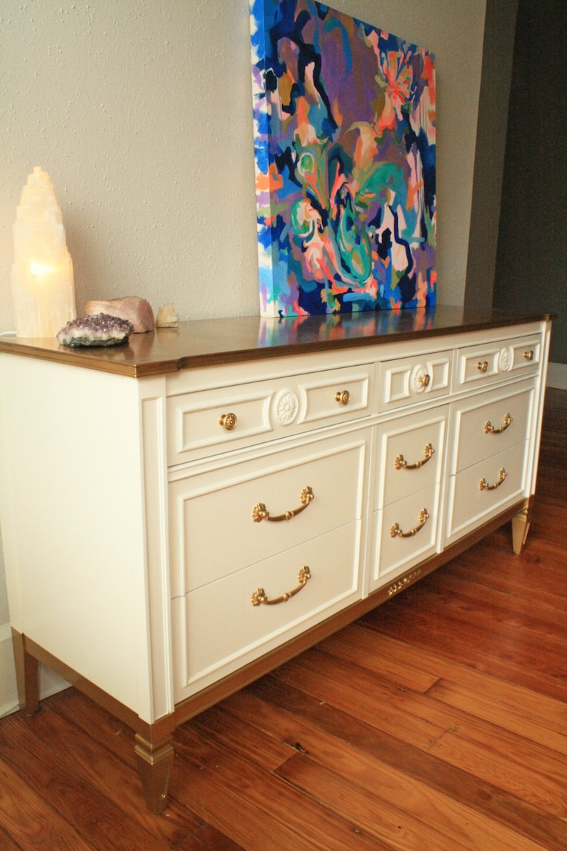 May include: A white dresser with gold accents and six drawers. The dresser has gold handles and decorative details. The dresser is sitting on a wooden floor. An abstract painting hangs on the wall behind the dresser.