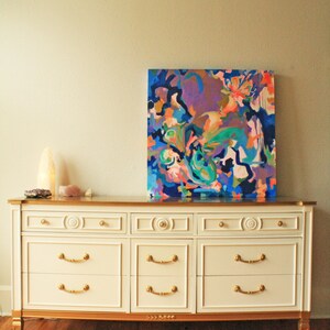 May include: A white dresser with six drawers and gold accents. The dresser has a gold top and gold trim around the drawers. The dresser is sitting on a wooden floor. A colorful abstract painting is hanging on the wall behind the dresser.
