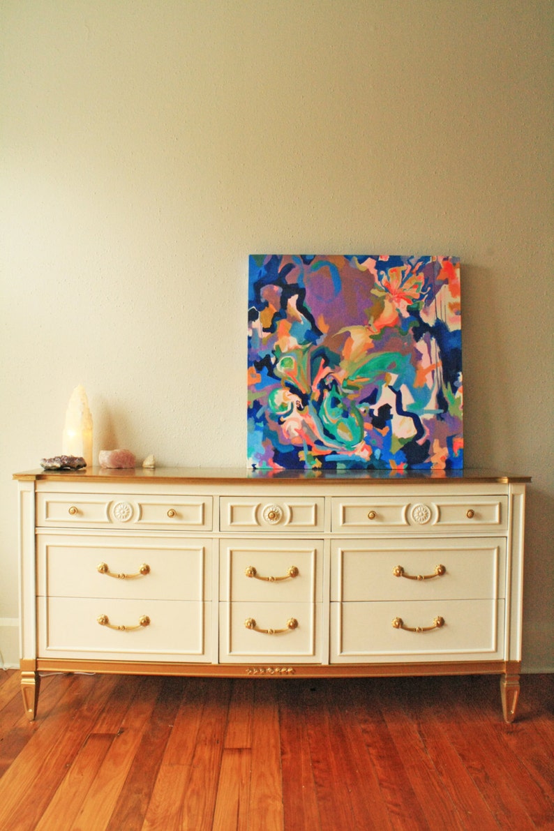 May include: A white dresser with six drawers and gold accents. The dresser has a gold top and gold trim around the drawers. The dresser is sitting on a wooden floor. A colorful abstract painting is hanging on the wall behind the dresser.
