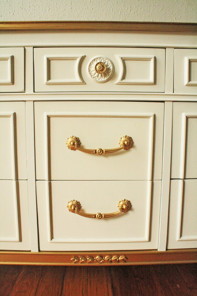 May include: White dresser with gold trim and ornate hardware. The dresser has six drawers with gold handles and a gold trim around the bottom.