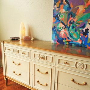 May include: A white dresser with gold accents and six drawers. The dresser has a gold top and sits on a wooden floor. An abstract painting hangs on the wall behind the dresser.