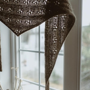 May include: A dark brown, triangular shawl with a delicate, openwork floral pattern. The knitted shawl is hanging near a window, showcasing its intricate design and texture. The shawl's edges are slightly curled.