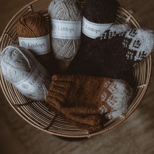 May include: Four balls of yarn in a wicker basket, two brown, one gray, and one black. The yarn is labeled "Lettlopi" and the basket contains two knitted items, one brown and one gray.