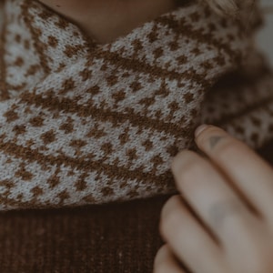 Könnte beinhalten: Nahaufnahme einer braunen und weißen Strickmütze mit geometrischem Muster. Die Mütze ist gefaltet und über die Schulter einer Person gelegt.