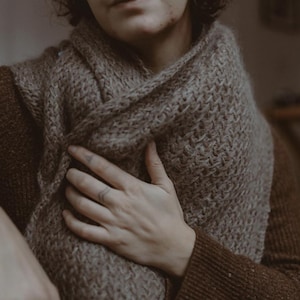 May include: A person wearing a brown knitted scarf with a textured pattern. The scarf is wrapped around the person's neck and chest, and the person is holding the scarf with one hand.