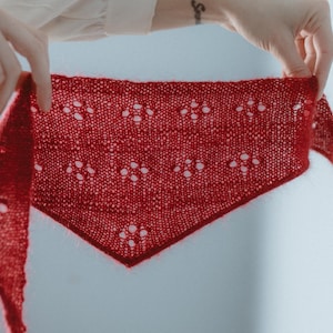 Op de afbeelding: Een rode gebreide sjaal met een delicaat kantpatroon.