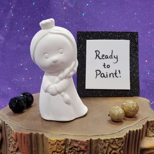 May include: White ceramic figurine of a girl with a crown, holding a paintbrush, ready to be painted. The figurine is on a wooden stump with gold glitter balls and a sign that says "Ready to Paint!"