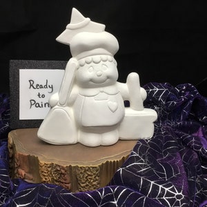 May include: White ceramic figurine of a witch holding a broom and a cauldron. The figurine is ready to be painted. The text "Ready to Paint" is written on a black card behind the figurine.