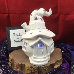 May include: A white ceramic house-shaped decoration with a witch hat design, featuring purple star accents. The house has a door and windows that emit a blue glow. The text "Ready to Paint!" is on a small sign.