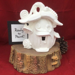 May include: A white ceramic fairy house, ready to paint, with whimsical details like a curly roof and mushroom accents. The house sits on a faux wood stump base. The sign says "Ready to Paint!"