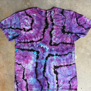 Pode incluir: Uma camiseta tie-dye em tons de roxo, azul e preto. A camisa apresenta gola redonda e mangas curtas. O padrão de tingimento tem um design linear rachado. A camisa é feita de um material macio.