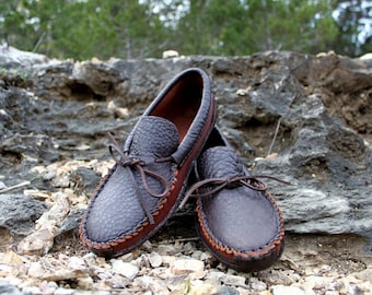 May include: A pair of dark brown leather moccasin-style shoes with a textured surface. The shoes have brown laces and stitching around the edges. The interior is a lighter brown color. The shoes are resting on a rocky surface.