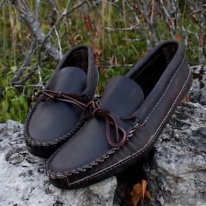 May include: A pair of dark brown leather moccasins with decorative stitching around the edge. The moccasins have laces that tie at the top.