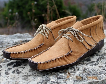 Puede incluir: Un par de mocasines de ante marrón con detalles de flecos y cordones. Los mocasines tienen una suela de cuero marrón y están sobre una roca.