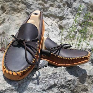 Peut inclure: Une paire de mocassins en cuir marron avec des coutures jaunes. Les mocassins ont une frange décorative et sont posés sur une surface rocheuse.