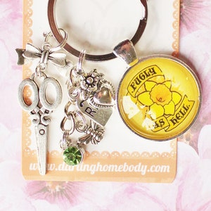 May include: A silver key chain with a charm that says "Ugly as hell" with a yellow daffodil on it. The charm is attached to a silver key ring with a silver scissor charm, a silver heart charm, and a green crystal charm.