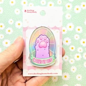 Op de afbeelding: Een roze en iriserende emaille pin met een kattenpoot ontwerp met de tekst "IN PAWS WE TRUST" op een groene banner. De pin is bevestigd aan een roze en witte achtergrondkaart met het Darling Homebody logo en website.