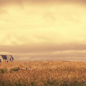 Könnte beinhalten: Ein kleines weißes Gebäude mit einem dunklen Dach steht auf einem grasbewachsenen Hügel vor einem bewölkten, gelben Himmel.