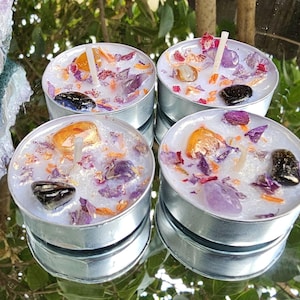 May include: Four tealight candles with a white wax base and various dried flowers and crystals embedded in the wax. The candles are in silver holders.