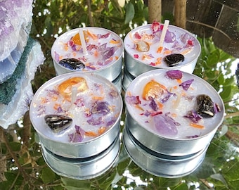 May include: Four tealight candles with a white wax base and various dried flowers and crystals embedded in the wax. The candles are in silver holders.