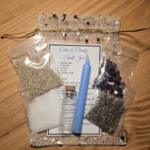May include: A clear, star-printed bag containing a spell jar kit. The kit includes a blue candle, amethyst chips, chamomile, lavender, sea salt, and a small glass bottle. The label reads "Calm & Clarity Spell Jar."