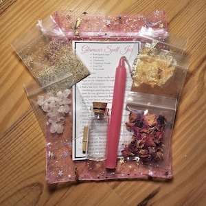 May include: A pink, star-speckled pouch containing a glamour spell kit. The kit includes a pink candle, rose quartz chips, rose petals, chamomile, cinnamon powder, gold foil, and a small glass bottle. The text "Glamour Spell Jar" is visible.