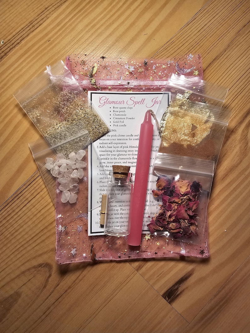 May include: A pink, star-speckled pouch containing a glamour spell kit. The kit includes a pink candle, rose quartz chips, rose petals, chamomile, cinnamon powder, gold foil, and a small glass bottle. The text "Glamour Spell Jar" is visible.