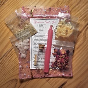 May include: A Glamour Spell Jar kit with a pink candle, rose quartz chips, rose petals, chamomile, cinnamon powder, and gold foil. The kit includes a small glass bottle and a pink organza bag with star accents. A card with instructions is also included.