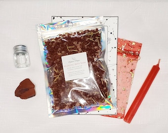 Puede incluir: Un baño de inmersión rojo con romero, una vela roja, una pequeña botella de vidrio transparente con tapa plateada y una piedra roja. El baño de inmersión está etiquetado como "Releasing Bath Soak Ritual Bath Soak for Cleansing, Transformation, and Letting Go".