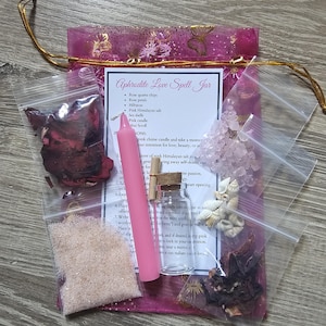 May include: A pink organza bag filled with various ingredients for a love spell, including rose quartz chips, rose petals, hibiscus, pink Himalayan salt, seashells, a pink candle, and a mini scroll. The bag is labeled "Aphrodite Love Spell Jar".