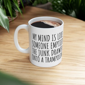 May include: White ceramic coffee mug with a black handle. The mug has black text that reads "My mind is like someone emptied the junk drawer onto a trampoline."