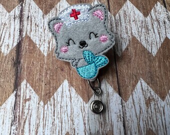 May include: A gray felt cat with a red cross on its hat and a blue mermaid tail. The cat is holding the tail in its paws. The cat is attached to a retractable badge holder.