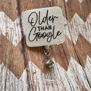 May include: White glitter badge holder with a black embroidered design that says "Older Than Google".