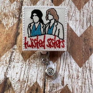 May include: White glitter badge holder with a white background and two cartoon figures of women with black hair and blue shirts. The text "Twisted Sisters" is in red letters below the figures.
