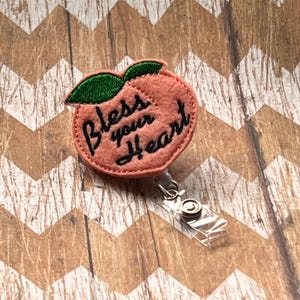 May include: Peach-shaped felt badge holder with a green leaf and the text "Bless your heart" in black embroidery. The badge holder is attached to a clear plastic retractable badge reel.