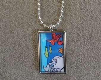 Op de afbeelding: Een zilveren ketting met een rechthoekige hanger met een cartoon afbeelding van Ursula uit De Kleine Zeemeermin. De hanger toont Ursula met haar tentakels, een rode krab en groene vissen.