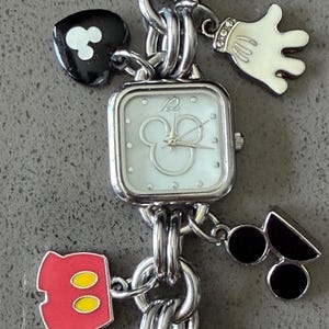 May include: Silver-toned charm bracelet watch featuring Mickey Mouse-themed charms. The watch has a square face with a Mickey Mouse silhouette. Charms include a Mickey glove, shorts, and a key. The bracelet is made of linked metal.