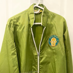 May include: A green satin jacket with a white zipper and white piping. The jacket has a patch on the left chest with a cartoon character and the text "Stay Gold".