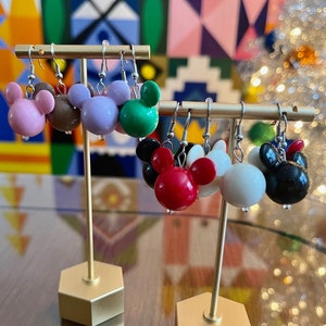 Puede incluir: Seis pares de pendientes colgantes con un dise&ntilde;o de cabeza de Mickey Mouse. Los pendientes est&aacute;n hechos de pl&aacute;stico y vienen en una variedad de colores, incluyendo rosa, marr&oacute;n, morado, verde, rojo, blanco y negro. Los pendientes se muestran en un soporte dorado.