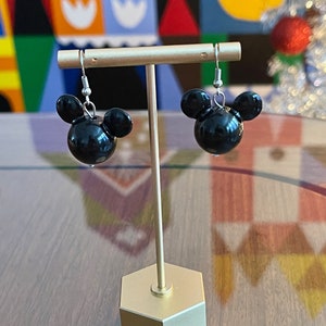 Puede incluir: Un par de pendientes negros con forma de Mickey Mouse con ganchos plateados. Los pendientes se muestran en un soporte dorado.