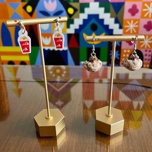 May include: Two pairs of earrings displayed on a gold stand. One pair features a red and white popcorn bucket charm, the other pair features a brown popcorn charm.