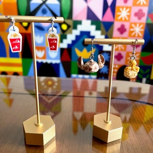 May include: Two pairs of earrings displayed on a gold stand. The earrings on the left are white with red accents and feature a popcorn bucket design. The earrings on the right are gold and feature a Stitch character from Lilo & Stitch.